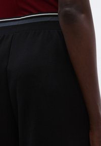 Czarne spodnie sportowe o fakturowanej powierzchni, z szerokim elastycznym pasem w talii ozdobionym czerwonymi, szarymi i białymi paskami u góry.