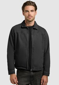 Hombre con una chaqueta negra con cremallera sobre una camiseta negra, posando frente a un fondo liso gris claro.