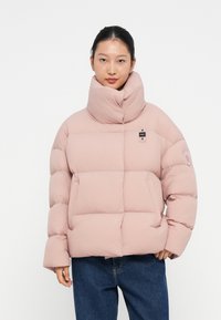 Ružová puff jacket s vysokým golierom, s štvorcovým prešívaným vzorom a nášivkou s logom na hrudi. Nosená nad džínsovými nohavicami.