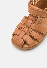 Braune Leder-Sandale mit geschlossenem Zehenbereich, ausgestattet mit geflochtenen Riemen und einem verstellbaren Klettverschluss. Glatte Innenauskleidung und flacher Sohle.