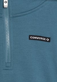 Sudadera con cremallera en color verde azulado, hecha de un tejido suave, que presenta un parche del logotipo "CONVERSE" en negro en contraste y un detalle de cremallera en cromado.