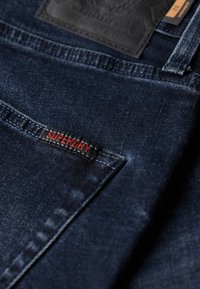 Jeans denim w ciemnym indygo o teksturowanym wykończeniu. Posiada czarną skórzaną naszywkę oraz logo "SUPERDRY" w czerwonym haftowanym napisie.