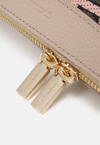 Gros plan d'une pochette en cuir beige avec doubles tirettes de fermeture éclair métalliques dorées gravées du nom de la marque Coccinelle.