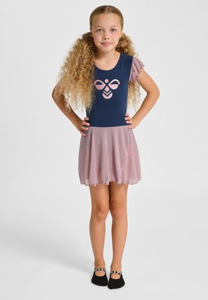 Ung pige med langt krøllet hår står med hænderne på hofterne iført en mørkeblå og pink kjole med en glitrende nederdel og sorte balletflade sko.