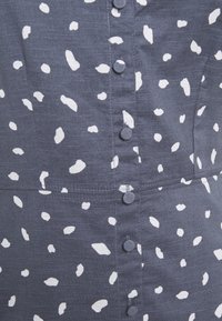 Szary materiał z białymi abstrakcyjnymi plamami, z zapinaniem na guziki. Gładka tekstura z widocznymi szwami i fałdami materiału.