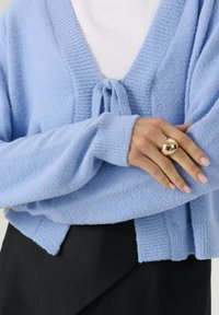 Cardigan a maglia blu chiaro con dettagli a cravatta indossato su un top bianco, abbinato a pantaloni neri, mostrando una mano con un grande anello d'oro e unghie rosa chiaro.