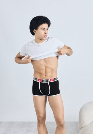 Zwarte boxershorts met witte accenten en een gebrandmerkte tailleband. Model draagt een effen wit T-shirt en trekt deze omhoog. Eenvoudige achtergrond.