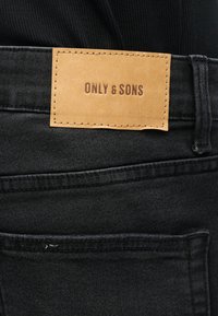 Ετικέτα από ταμπά δέρμα με χαραγμένο το "ONLY & SONS" στη μέση ενός μαύρου τζιν με εμφανείς ραφές και λεπτομέρειες στο πίσω τσέπη.