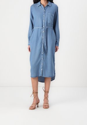 Kvinde iført en langærmet, knap-down blå denim kjole med bælte i taljen og tan stroppet højhælede sandaler står på en hvid gulv.