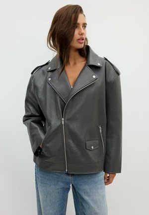 Kvinde iført oversized sort læderjakke med sølvlynlåse og lyseblå jeans, ser til siden mod en ensfarvet grå baggrund.