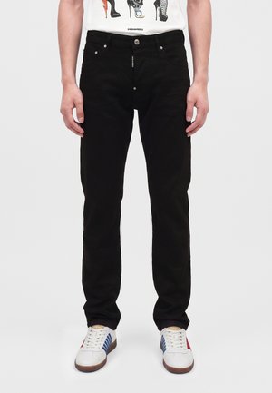 Uomo con jeans neri, t-shirt bianca con grafica e sneakers bianche con strisce blu e rosse, in piedi davanti a uno sfondo bianco.