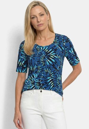 Vrouw met lang blond haar, gekleed in een blauw patroon kort mouwtopje en witte broek, staand tegen een effen lichte achtergrond.