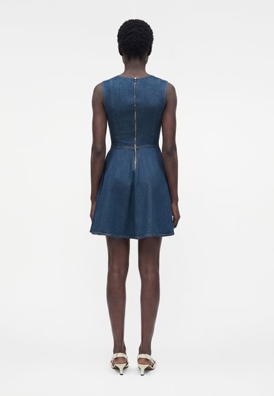 Γυναίκα που στέκεται πλάτη, φοράει μπλε τζιν φόρεμα χωρίς μανίκια με ορατό φερμουάρ στην πλάτη και μπεζ τακούνια σανδάλια.