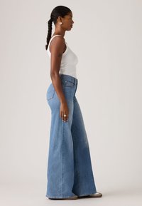 Φαρδιά τζιν παντελόνια σε ανοιχτό μπλε χρώμα με ψηλή μέση, φτιαγμένα από ντένιμ. Διαθέτουν μπροστινές τσέπες και ξεθωριασμένο εφέ. Φοριούνται με λευκό τοπ χωρίς μανίκια.