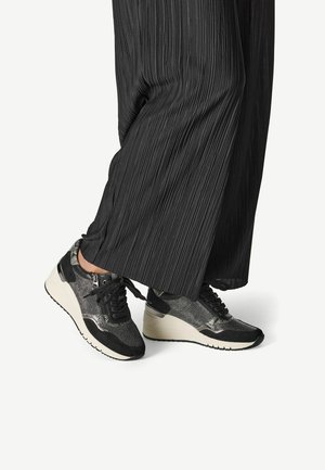 Sneaker neri e grigi con tomaia testurizzata, chiusura con lacci e suola bianca imbottita. Indossati con pantaloni ampi neri a pieghe.