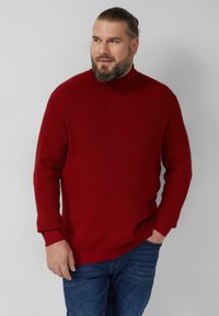 Czerwony prążkowany sweter z golfem i długimi rękawami, o fakturowanym wykończeniu i luźnym kroju, zestawiony z niebieskimi jeansami.