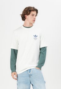 Homme adulte portant un t-shirt blanc à manches courtes avec un logo Adidas bleu et le texte "HEALTH & FITNESS Club", superposé sur une chemise à manches longues verte.