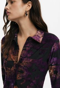 Maglietta nera a maniche lunghe con motivi astratti viola e marroni, colletto evidente e chiusura a bottone nella parte anteriore. Orecchini a forma di stella.