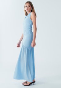 Gaiši zila maxi kleita ar pieguļošu augšdaļu un savāktu tekstūru, kas plūst vaļējā svārkā. Valkāta ar melnām čībām.