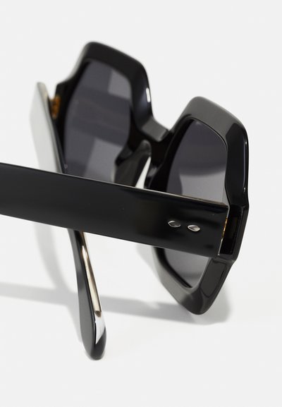 Occhiali da sole oversize neri con una montatura geometrica audace, lenti scure e aste nere eleganti. Presentano una finitura lucida e sottili accenti metallici.