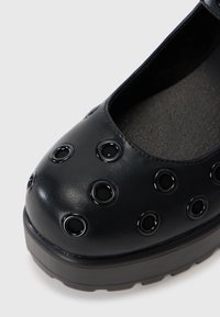 Black leather shoe featuring a rounded toe, textured surface, and decorative circular cutouts with glossy black accents. Flat sole design.