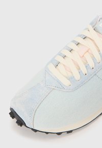 Lys blå sneaker med et struktureret kanvasoverdel, cremefarvede snørebånd og en sort gummisål med trækkenspidser. Glatte pastelfarvede accenter er synlige.