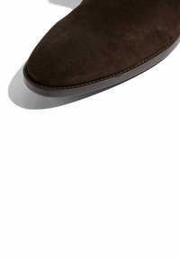 Zapato de ante marrón con punta afilada, detalles de costura a lo largo del borde y una suela plana. La textura parece suave y lisa.