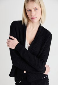 Cardigan negru cu model texturat, cu o croială lejeră, deschis în față și mâneci lungi. Textura este tricotată, sporind căldura și confortul.
