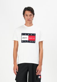 Homme portant un T-shirt blanc Tommy Hilfiger avec le logo des États-Unis et de l'équipe Sail GP, associé à un short noir arborant un patch en forme de drapeau.