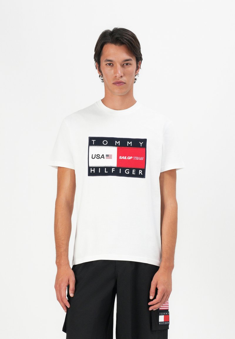 Homme portant un T-shirt blanc Tommy Hilfiger avec le logo des États-Unis et de l'équipe Sail GP, associé à un short noir arborant un patch en forme de drapeau.