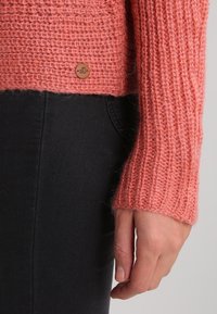 Pull en laine douce de couleur corail clair avec un côtes texturé. Il présente un ourlet court et une discrète étiquette logo en cuir.