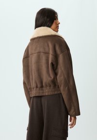 Giacca corta in finta suede marrone con colletto in shearling crema, vita elasticizzata e texture liscia. Presenta una vestibilità comoda e dettagli sulle cuciture laterali.