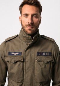 Giacca militare verde oliva con colletto alto, due tasche sul petto e toppe di tessuto blu con ali e testo J.P. 18-80.