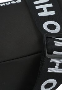 Borsa di stoffa nera con un logo e testo bianchi ben visibili sulla tracolla, con una texture liscia e forma rettangolare. Inclusa una tracolla regolabile.