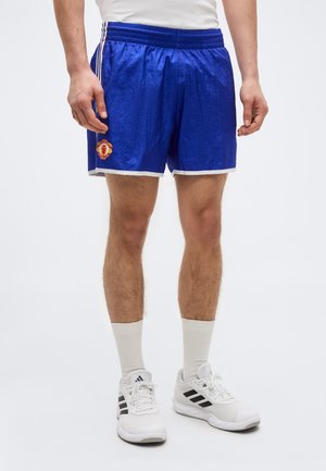 Muškarac u plavim sportskim kratkim hlačama s logom Manchester Uniteda, bijelim čarapama i bijelim Adidas tenisicama, stoji na jednobojnoj pozadini.