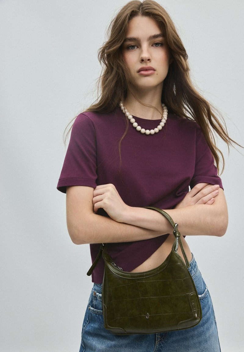 Jeune femme aux longs cheveux bruns porte un haut violet, un collier de perles, un jean bleu et tient un sac à main vert les bras croisés.