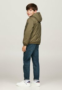 Giacca con cappuccio verde oliva con polsini elasticizzati, jeans in denim blu e sneakers bianche con accenti navy e rossi. Tessuto texturizzato e vestibilità regolare.