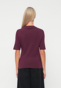 Femme aux longs cheveux blonds portant un haut prune à manches courtes et un pantalon noir, se tenant face à un fond clair uni.