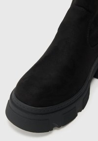 Črni gležnjarji iz semiša s debelim črnim gumijastim podplatom. Imajo gladko teksturo, zaobljen prst in subtilne detajle šivov.