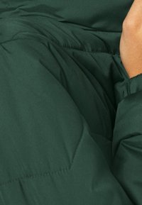 Giacca puffer verde scuro caratterizzata da una texture liscia, design trapuntato e polsini elasticizzati arricciati. Una leggera lucentezza aggiunge profondità alla superficie.