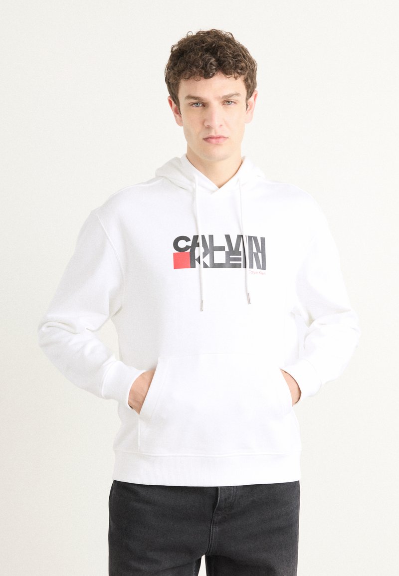 Fehér kapucnis pulóver elülső zsebbel, fekete és piros "CALVIN KLEIN" logóval. Az anyag puhanak tűnik, húzózsinóros részletekkel.