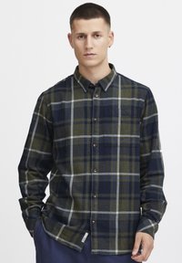Camisa de franela en un patrón de cuadros en azul marino, verde y gris. Cuenta con un cuello de botón, bolsillo frontal y cierre de botones. Textura suave.