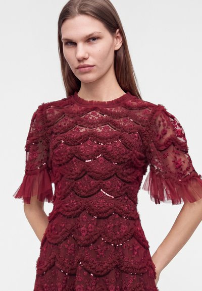 Jeune femme aux cheveux châtain clair et raides, portant une robe en dentelle bordeaux avec des motifs floraux et des manches bouffantes transparentes.