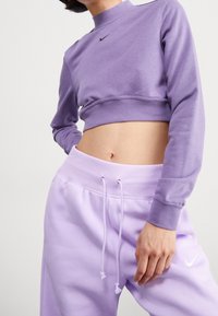 Felpa corta viola con collo alto, maniche lunghe e logo nero. Abbinata a pantaloni jogger lavanda coordinati con coulisse e logo.