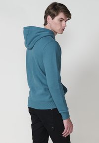 Felpa blu con cappuccio, dotata di polsini e orlo a coste, vestibilità ampia e una texture liscia. Il modello si sta allontanando dalla fotocamera.