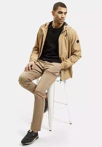 Beżowa, lekka kurtka z kapturem, czarna bluza pod spodem i beżowe, dopasowane spodnie. Czarne buty sportowe i okrąły detal z logo na kurtce.