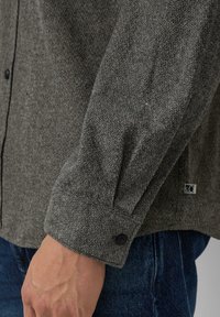 Grijze gestructureerde button-up shirt met zwarte knopen, met een ontspannen pasvorm en een klein logo-etiket aan de zijkant. Close-up van het mouwdetail weergegeven.