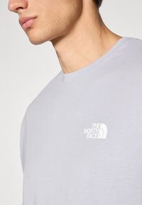 Šviesiai pilkas trumpomis rankovėmis marškinėliai, pagaminti iš minkštos medžiagos. Yra mažas baltas "The North Face" logotipas kairėje krūtinės pusėje.