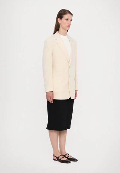 Blazer in lino crema con un singolo bottone, abbinato a pantaloni corti neri fino al ginocchio e scarpe nere con tacco e dettagli a fascia.