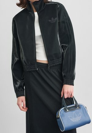 Kvinde iført en sort lynlåstrøje og nederdel, der holder en lille blå håndtaske med et hvidt logo.